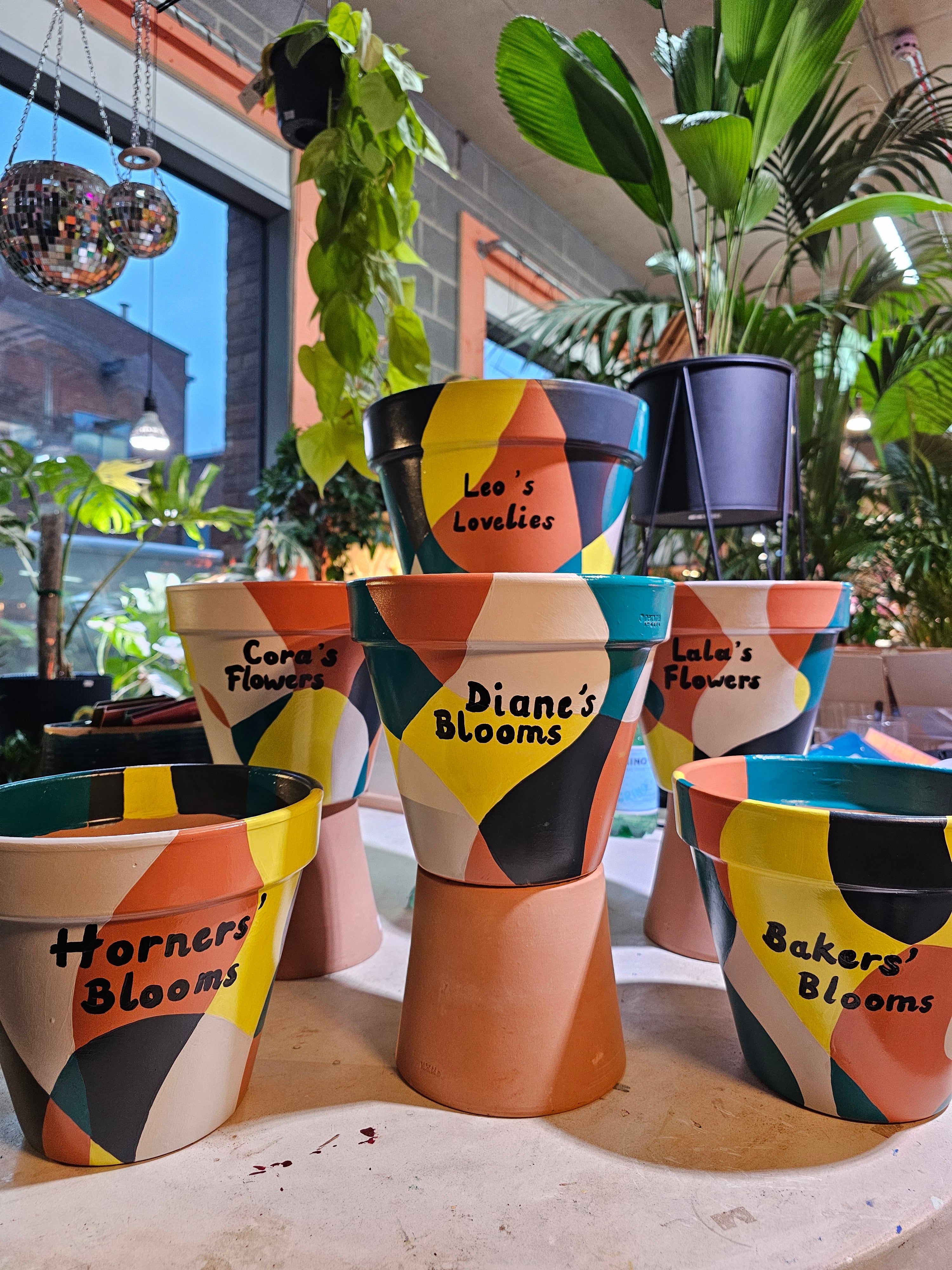 Colorful flower pots with personalised names hand-painted, surrounded by plants.